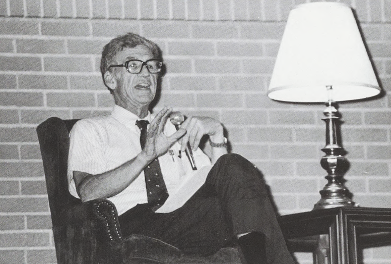 Black and white photo of Chancellor William H. Danforth speaking into a microphone while sitting in a chair next to a table with a lamp on it.