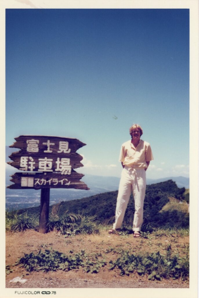 Color photo of WashU alumnus Kevin Crowell standing next to a sign with Japanese writing on it in a mountainous region of Japan on a sunny day in the 1970s.